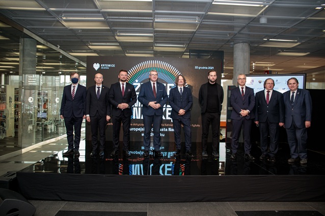 Dziewięciu mężczyzn w formalnych strojach stoi w rzędzie w pomieszczeniu, pozując do grupowego zdjęcia przed dużym ekranem cyfrowym z polskim tekstem i okrągłą grafiką. Jeden mężczyzna ma na sobie maskę. Sceneria wydaje się nowoczesna i profesjonalna.