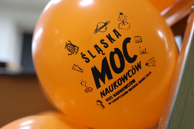 Pomarańczowy balonik z czarnymi ilustracjami ikon naukowych i polskim tekstem: Śląska Moc Naukowców. Noc Naukowców w Europejskim Mieście Nauki 2024.