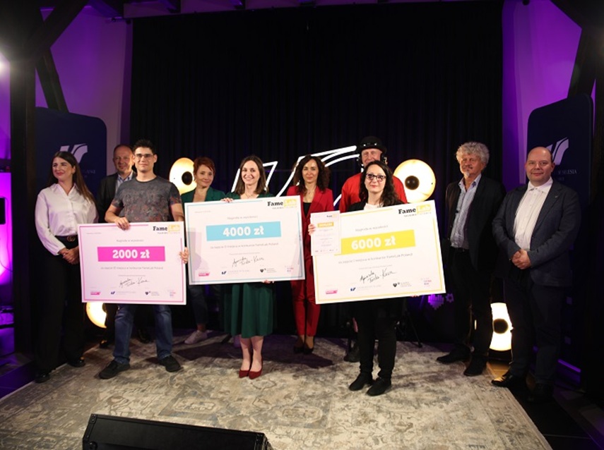 Grupa dziewięciu osób stoi na wyłożonej dywanem scenie, trzy z nich trzymają duże czeki z nagrodami na 2000, 4000 i 6000 złotych. Tło jest ciemne, z włączonymi światłami scenicznymi i banerami wydarzeń po obu stronach.
