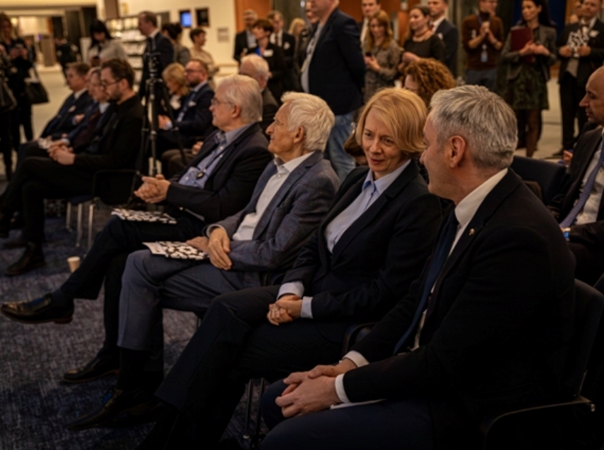 Grupa ludzi w formalnych strojach siedzi blisko siebie w rzędach na imprezie w pomieszczeniu, uważnie słuchając i rozmawiając. Inni stoją w tle przy stołach i ekspozycjach.