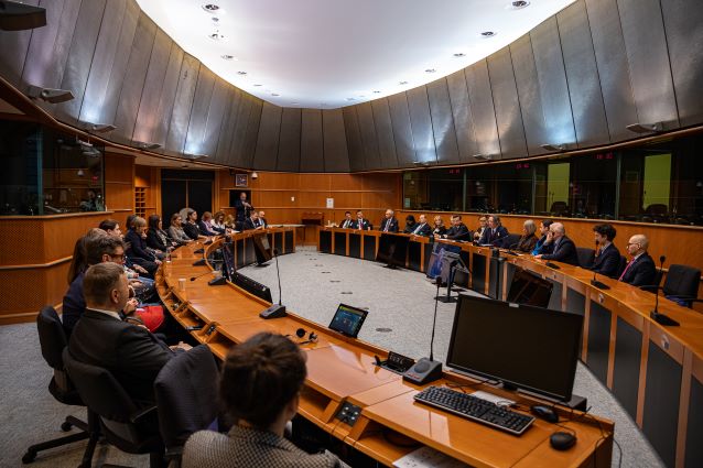 Duża sala konferencyjna z ludźmi siedzącymi wokół zakrzywionego, drewnianego stołu, zaangażowanymi w formalne spotkanie lub dyskusję. Na stołach widoczne są monitory komputerowe i mikrofony.