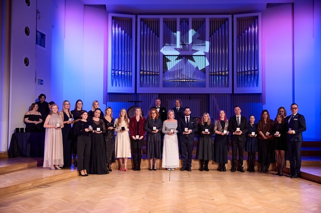 Grupa formalnie ubranych osób stoi na scenie, trzymając nagrody, pozuje do zdjęcia na tle wielkich piszczałek organowych i oświetlonego, geometrycznego tła w przestronnej, eleganckiej sali.