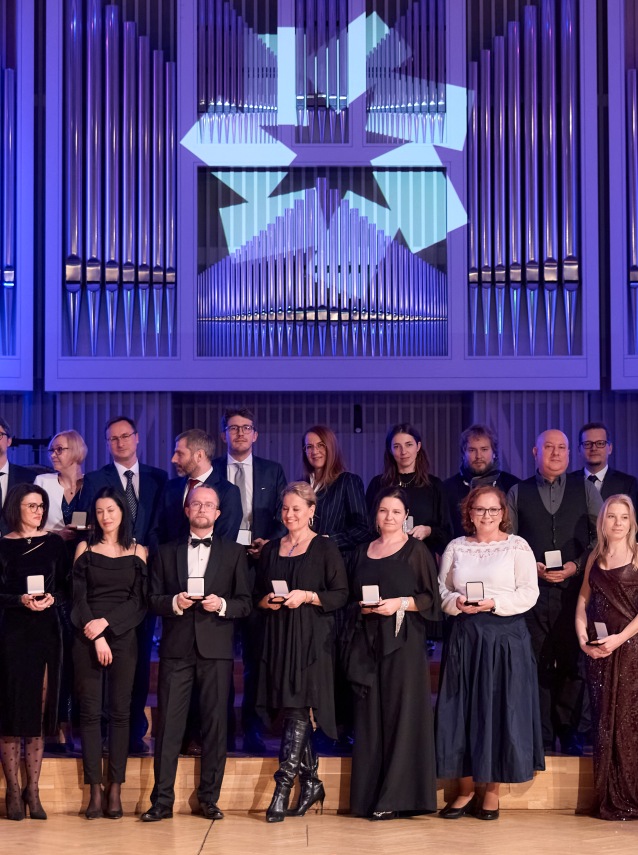 Grupa formalnie ubranych osób stoi na scenie, trzymając medale. W tle widać duże organy piszczałkowe i geometryczną dekorację świetlną.