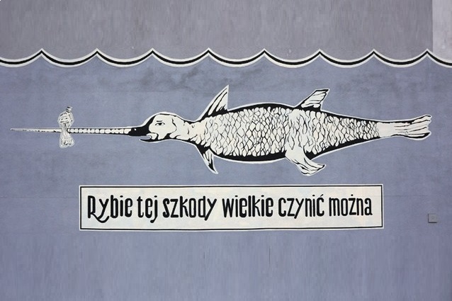 Malowidło ścienne przedstawiające dużą rybę z długim nosem zwróconą w stronę małej ryby, namalowane na niebieskiej ścianie. Pod rybą znajduje się polska fraza: Rybie tej szkody wielkie czynić można.