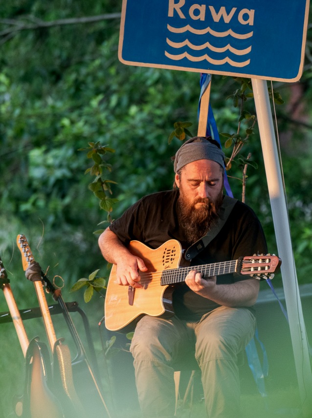Brodaty mężczyzna w chuście na głowie gra na gitarze akustycznej na zewnątrz, siedząc przy niebieskim znaku „Rawa”. Dwie inne gitary leżą nieopodal, a w tle widać zielone liście.