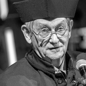Starszy mężczyzna w okularach i tradycyjnej czapce akademickiej i todze mówi do mikrofonu. Obraz jest czarno-biały.