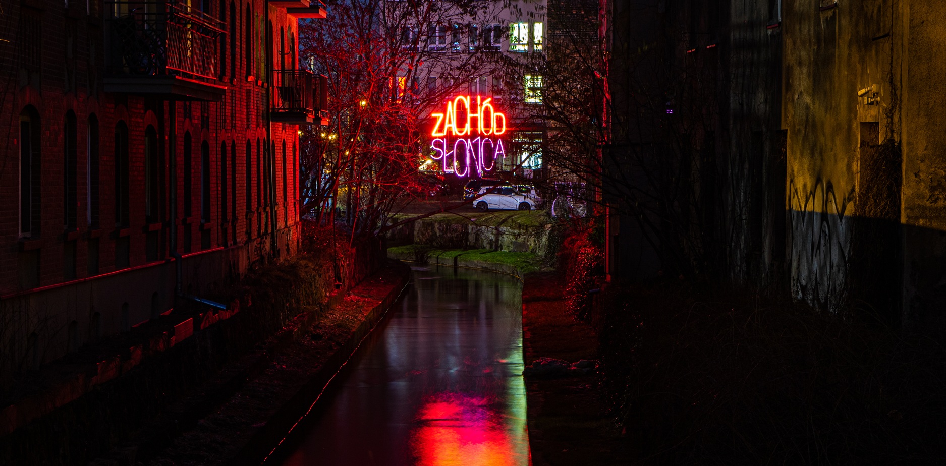 W nocy między budynkami przebiega wąski kanał, oświetlony refleksami czerwonych neonów i świecącym neonowym napisem ZAKUS SLAMO w oddali, obok zaparkowanego białego samochodu.