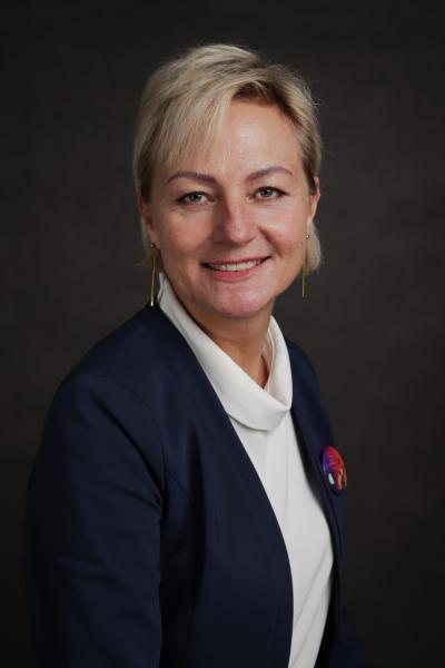 Kobieta z krótkimi blond włosami, ubrana w ciemny blezer na białym topie, uśmiecha się do kamery na ciemnym tle. Nosi długie kolczyki i kolorową broszkę na blezerze.