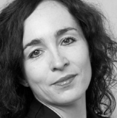 Czarno-biały portret kobiety o falowanych ciemnych włosach, jasnej karnacji i delikatnym uśmiechu, ubranej w ciemną bluzkę i patrzącej prosto w obiektyw.