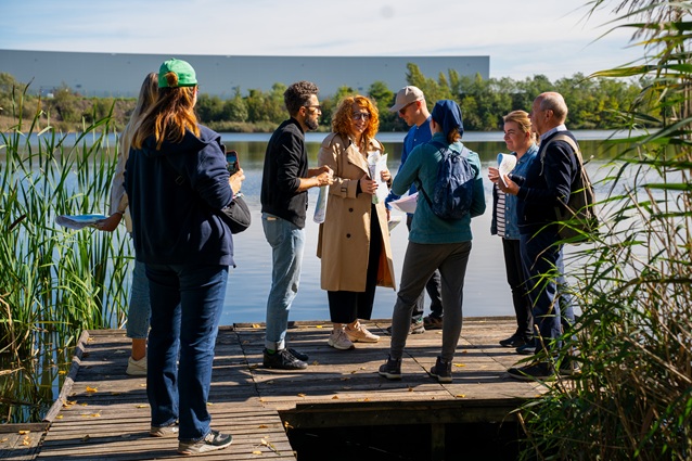 Grupa ludzi stoi na drewnianym pomoście nad jeziorem, trzyma papiery i rozmawia. Niektórzy noszą codzienne ubrania i plecaki, a w pobliżu rosną trzciny. W tle widać drzewa i duży budynek.