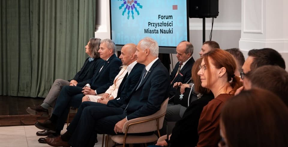 Kilka osób w formalnych strojach siedzi w rzędzie, uważnie zwróconych do przodu na imprezie wewnętrznej. Za nimi znajduje się ekran wyświetlający tekst Forum Przyszłości Miasta Nauki. Atmosfera wydaje się profesjonalna i poważna.