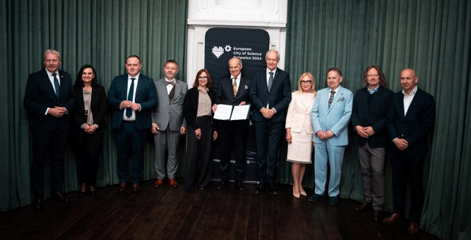 Grupa jedenastu formalnie ubranych osób stoi w rzędzie na ciemnej drewnianej podłodze, pozując do zdjęcia przed zielonymi zasłonami i banerem z napisem Europejskie Miasto Nauki 2024. Dwie osoby w środku trzymają certyfikat.