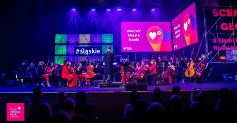 Orkiestra symfoniczna występuje na scenie oświetlonej żywymi fioletowymi i niebieskimi światłami. Duże ekrany wyświetlają "Wieczór Miasta Nauki" i "#śląskie". Członkowie publiczności są widoczni w sylwetkach.