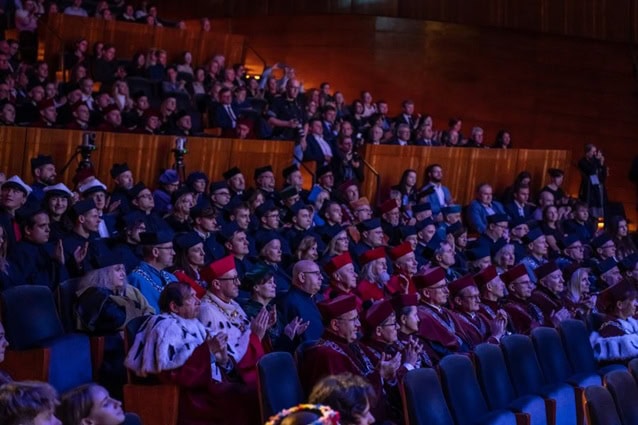 Duża publiczność, wielu ubranych w akademickie togi i czapki, siedzi w słabo oświetlonym audytorium, uważnie obserwując wydarzenie na scenie. Niektórzy klaszczą, a siedzenia są ustawione w rzędach.