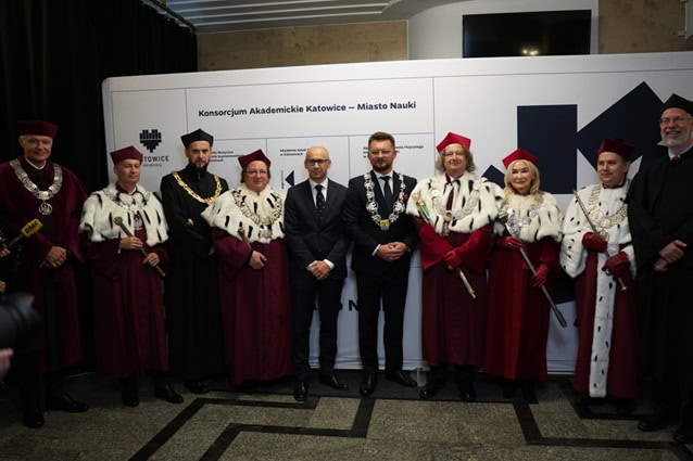 Grupa rektorów i przedstawicieli władz miasta w uroczystych szatach i łańcuchach stoi z dwoma mężczyznami w garniturach przed białym tłem z logo i tekstem, pozując do oficjalnego zdjęcia na imprezie wewnętrznej.