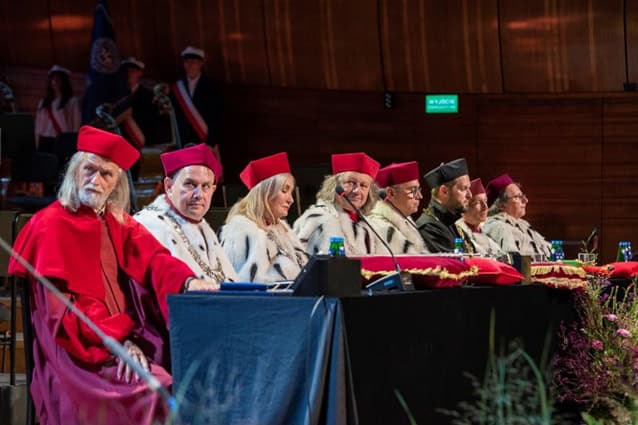 Grupa rektorów akademickich w czerwono-białych szatach ceremonialnych i kapeluszach siedzi przy długim stole na scenie podczas oficjalnego wydarzenia uniwersyteckiego w wyłożonym drewnianymi panelami audytorium.