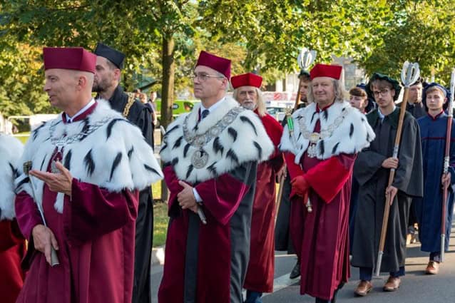 Grupa rektorów akademickich w czerwonych szatach z białymi futrzanymi wykończeniami i czerwonymi czapkami idzie w procesji na zewnątrz; inni uczestnicy w różnych akademickich regaliach idą za nimi. W tle widoczne są drzewa i światło słoneczne.