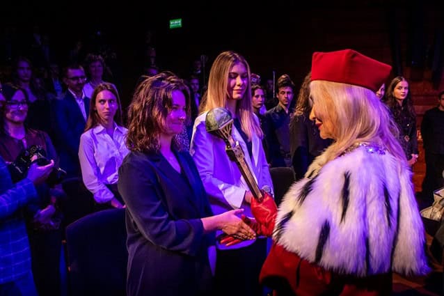 Kobieta w akademickiej szacie i czerwonym kapeluszu podaje rękę innej kobiecie w ciemnym garniturze, podczas gdy tłum ludzi obserwuje ją w słabo oświetlonym pomieszczeniu.