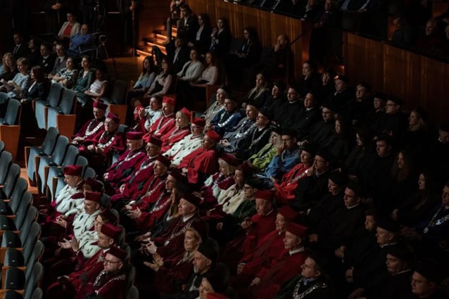 Grupa ludzi w akademickich szatach, niektórzy w czerwieni i bieli, inni w czerni, siedzą w rzędach podczas oficjalnej ceremonii w audytorium wypełnionym uczestnikami.