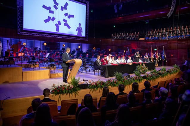 Uroczysta inauguracja w audytorium, z mówcą na podium, urzędnikami siedzącymi przy stole i dużym ekranem pokazującym logo. Członkowie publiczności obserwują z miejsc.