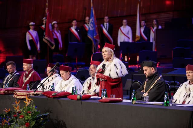 Grupa rektorów w akademickich szatach siedzi przy długim stole na scenie; kobieta stoi i mówi do mikrofonu. W tle flagi i formalnie ubrani ludzie.