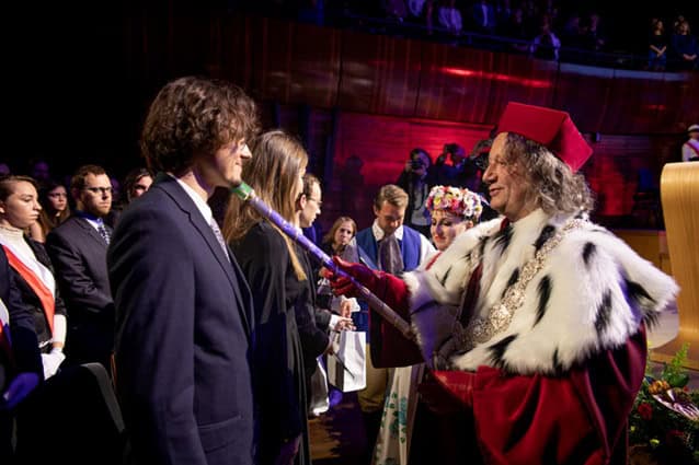 Mężczyzna w uroczystych szatach i kapeluszu dotyka berłem ramienia młodego mężczyzny podczas oficjalnej ceremonii, podczas gdy inni obserwują w tle. Otoczenie jest eleganckie i świąteczne.