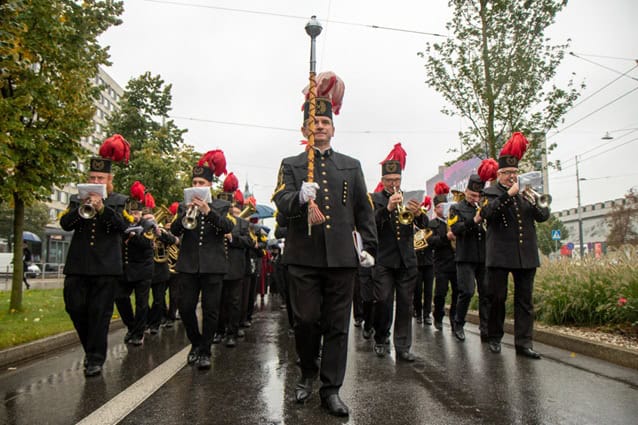 Orkiestra marszowa w czarnych mundurach z czerwonymi pióropuszami na czapkach paraduje mokrą ulicą, prowadzona przez pałkarza. Członkowie zespołu grają na instrumentach podczas spaceru na zewnątrz w pochmurny dzień.