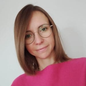 Kobieta z prostymi, sięgającymi ramion brązowymi włosami, w okularach i jasnoróżowym topie, uśmiecha się lekko, stojąc na jasnym tle.