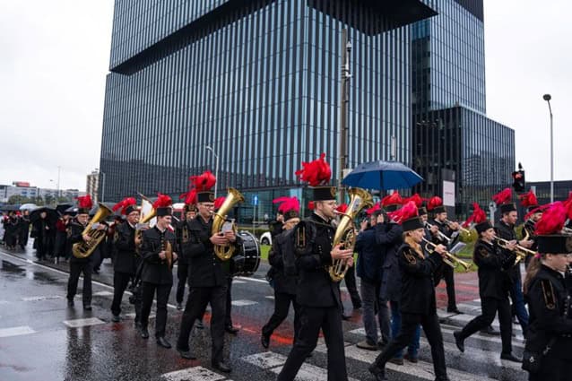 Orkiestra marszowa w czarnych mundurach z czerwonymi kapeluszami z piórami gra na instrumentach dętych, idąc ulicą miasta w pobliżu nowoczesnych szklanych budynków w deszczowy dzień; niektórzy ludzie niosą parasole.
