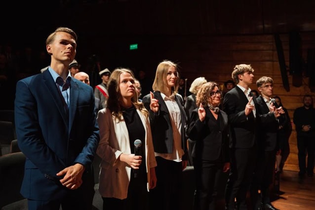 Grupa ludzi w formalnych strojach stoi w pomieszczeniu, niektórzy trzymają prawe ręce w górze, składając przysięgę. Jedna osoba z przodu trzyma mikrofon.