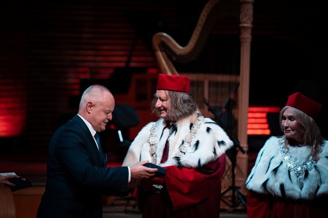 Mężczyzna w garniturze podaje rękę uśmiechniętemu naukowcowi w czerwonej czapce i futrzanej szacie podczas oficjalnej ceremonii, podczas gdy inny podobnie ubrany naukowiec stoi obok.