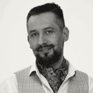 Czarno-biały portret mężczyzny z krótkimi włosami i brodą, ubranego w kamizelkę w kratę na białej koszuli i wzorzysty szalik zawiązany na szyi, uśmiechającego się lekko do kamery.