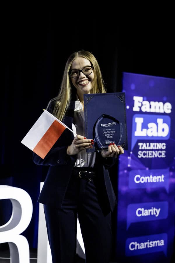 Uśmiechnięta kobieta w garniturze trzyma polską flagę i tabliczkę z nagrodą przed wyświetlaczem FameLab Talking Science, na którym widnieją takie słowa jak Content, Clarity i Charisma.