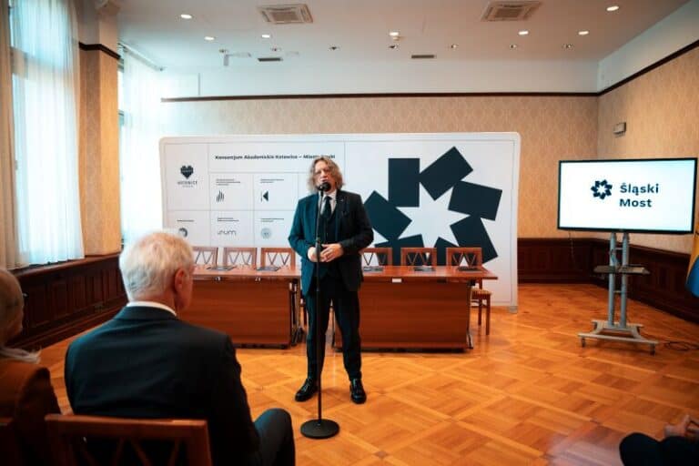 Mężczyzna w garniturze przemawia do mikrofonu w sali konferencyjnej z drewnianą podłogą. Za nim znajdują się puste krzesła, duże tło z logo i ekran wyświetlający "Śląski Most". Kilka osób siedzi i słucha.