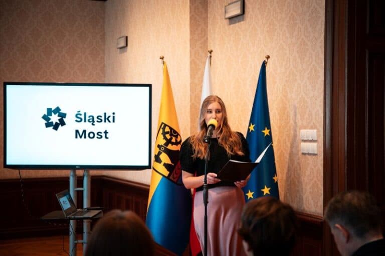 Kobieta stoi na podium i mówi do mikrofonu, trzymając w ręku dokumenty. Za nią znajdują się flagi Polski, Unii Europejskiej i żółto-czarna flaga. Na ekranie wyświetlane jest logo i napis Śląski Most. Na pierwszym planie siedzą ludzie.