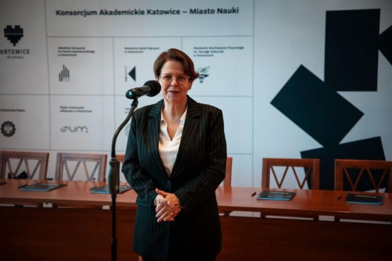 Kobieta w ciemnym garniturze w prążki stoi przy mikrofonie i przemawia. Za nią znajduje się stół z krzesłami i tłem wyświetlającym logo i tekst w języku polskim, nawiązujący do Katowic i konsorcjum akademickiego.