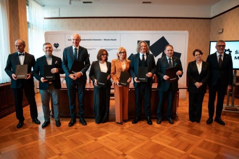 Osiem osób w formalnych strojach stoi w rzędzie w pomieszczeniu, trzymając czarne teczki i pozując do zdjęcia grupowego. Wydają się być na oficjalnym wydarzeniu lub ceremonii, z markowym tłem i wypolerowaną drewnianą podłogą.