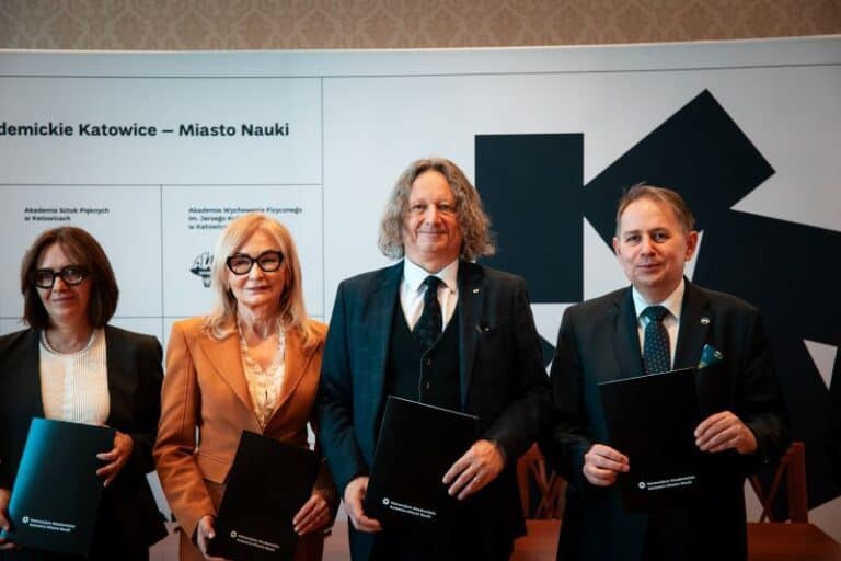 Cztery osoby w formalnych strojach stoją obok siebie, trzymając czarne teczki, pozując do zdjęcia przed ścianą z tekstem i geometrycznymi wzorami na oficjalnym wydarzeniu.