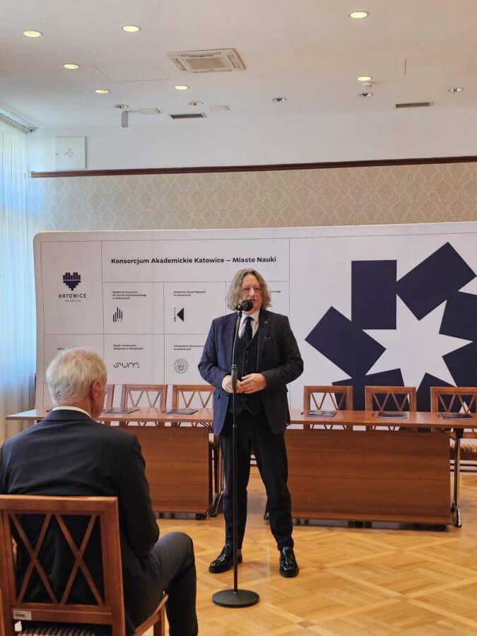 Rektor UŚ prof. dr hab. Ryszard Koziołek przemawia do mikrofonu w sali konferencyjnej, stojąc twarzą do członka publiczności. Za nim znajduje się wyświetlacz z wykresami i logo oraz kilka pustych krzeseł wzdłuż drewnianego stołu.