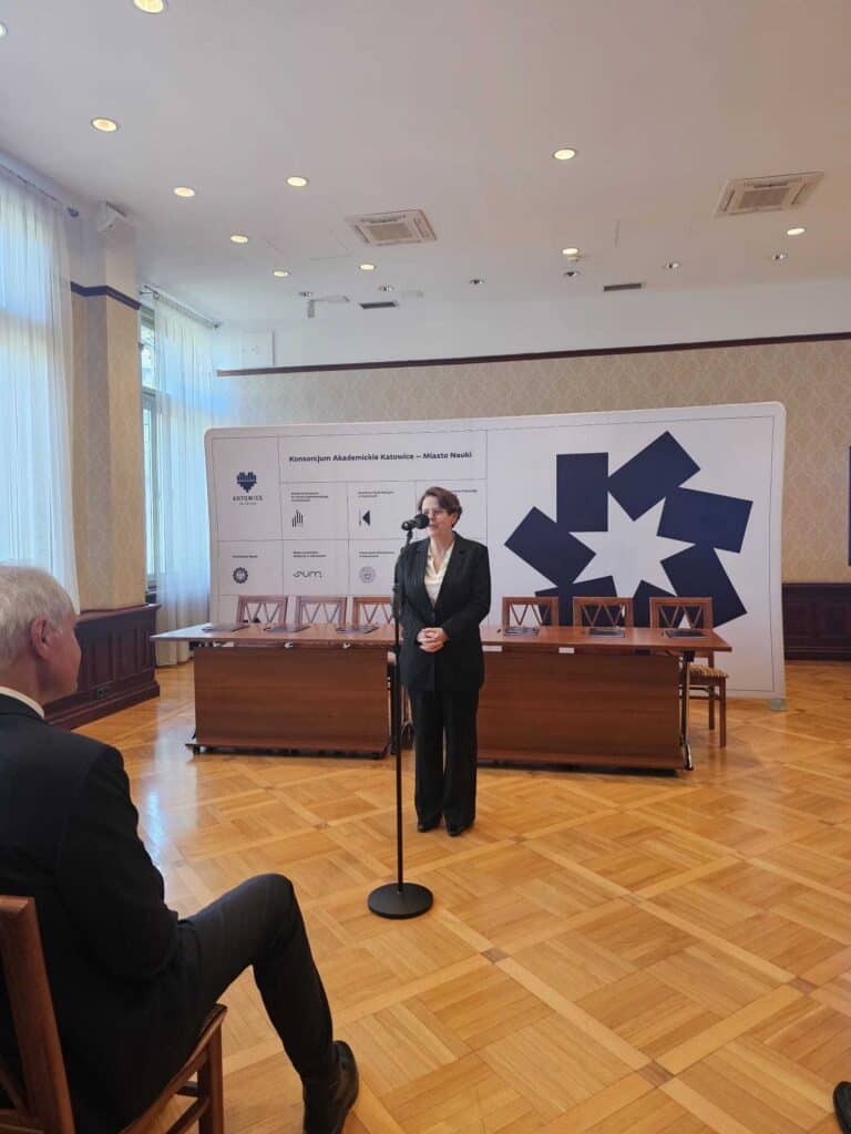 Kobieta w garniturze stoi i przemawia do mikrofonu w formalnym pomieszczeniu z drewnianą podłogą, podczas gdy siedzący mężczyzna słucha. Za nią znajduje się stół i baner z logo i geometrycznym niebieskim wzorem.