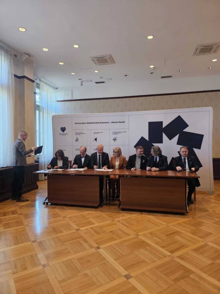 Siedem osób siedzi przy długim stole w sali konferencyjnej, podpisując dokumenty, podczas gdy jeden mężczyzna stoi z boku, trzymając dokumenty. Za nimi znajduje się biała tablica z logo i tekstem. Pokój ma drewniane podłogi i duże okna.