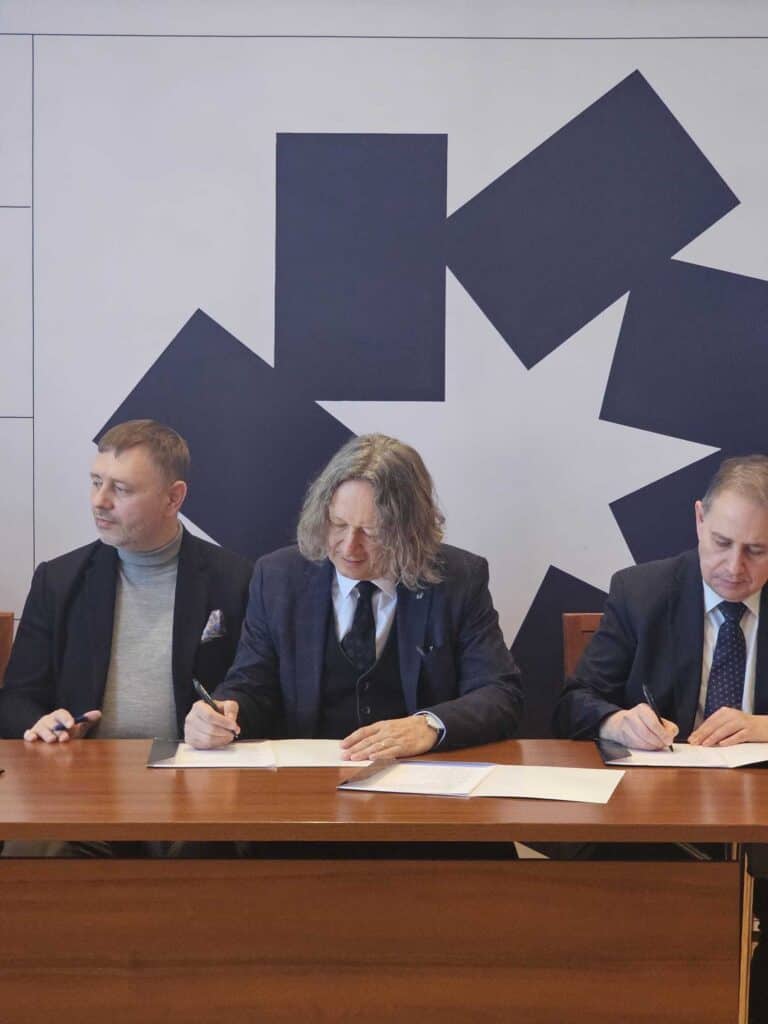 Trzech mężczyzn w garniturach siedzi przy stole i podpisuje dokumenty. Za nimi znajduje się ściana z dużym geometrycznym wzorem przedstawiającym ciemne kształty i białą gwiazdę pośrodku.